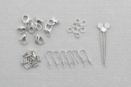"Assorted sterling silver jewelry findings including lobster claw clasps, jump rings, ear wires, crimp tubes, and head pins arranged on a linen texture background"